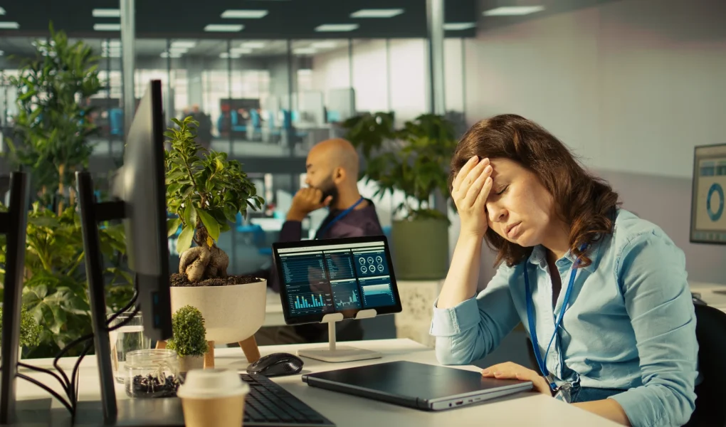 A stressed or exhausted businesswoman sitting at her office desk with her eyes closed and her hand pressed to her forehead. Her desk features a closed laptop, a tablet displaying data charts, and computer monitors, with a blurred colleague and office plants in the background.