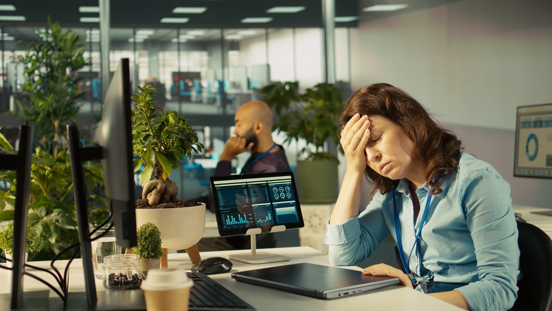 A stressed or exhausted businesswoman sitting at her office desk with her eyes closed and her hand pressed to her forehead. Her desk features a closed laptop, a tablet displaying data charts, and computer monitors, with a blurred colleague and office plants in the background.