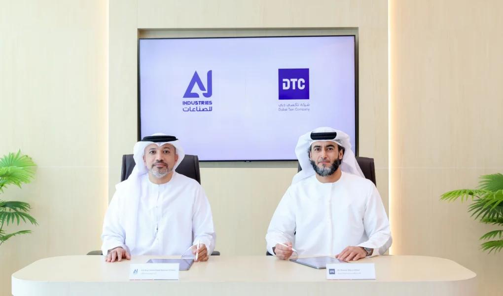 Two men wearing traditional Emirati attire (kandura and ghitra) seated at a modern desk for a partnership signing. A large display behind them shows the logos for AJ Industries and Dubai Taxi Company (DTC). Small nameplates sit on the table in front of each representative.