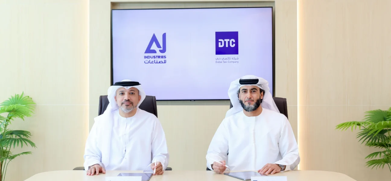 Two men wearing traditional Emirati attire (kandura and ghitra) seated at a modern desk for a partnership signing. A large display behind them shows the logos for AJ Industries and Dubai Taxi Company (DTC). Small nameplates sit on the table in front of each representative.
