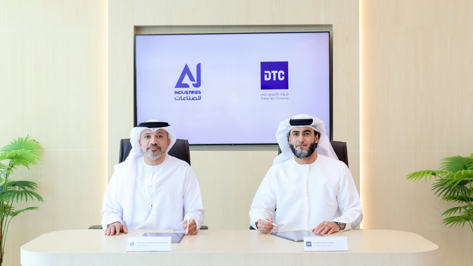 Two men wearing traditional Emirati attire (kandura and ghitra) seated at a modern desk for a partnership signing. A large display behind them shows the logos for AJ Industries and Dubai Taxi Company (DTC). Small nameplates sit on the table in front of each representative.