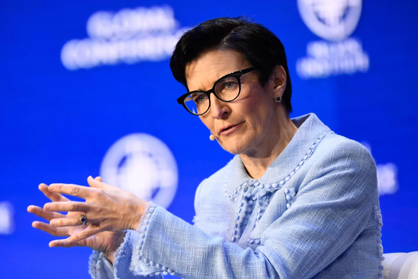 Jane Fraser, CEO of Citigroup, speaking during a panel discussion at a conference. She has short dark hair, wears black-rimmed glasses and a light blue textured jacket, gesturing with her hands against a bright blue background featuring blurred conference branding.