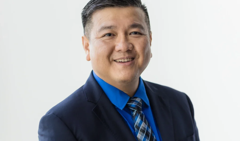 A professional headshot of Naaman Lee, an Asian man with short, styled dark hair, smiling warmly at the camera against a plain white background. He is wearing a dark navy suit jacket, a vibrant blue dress shirt, and a patterned blue and gray tie.