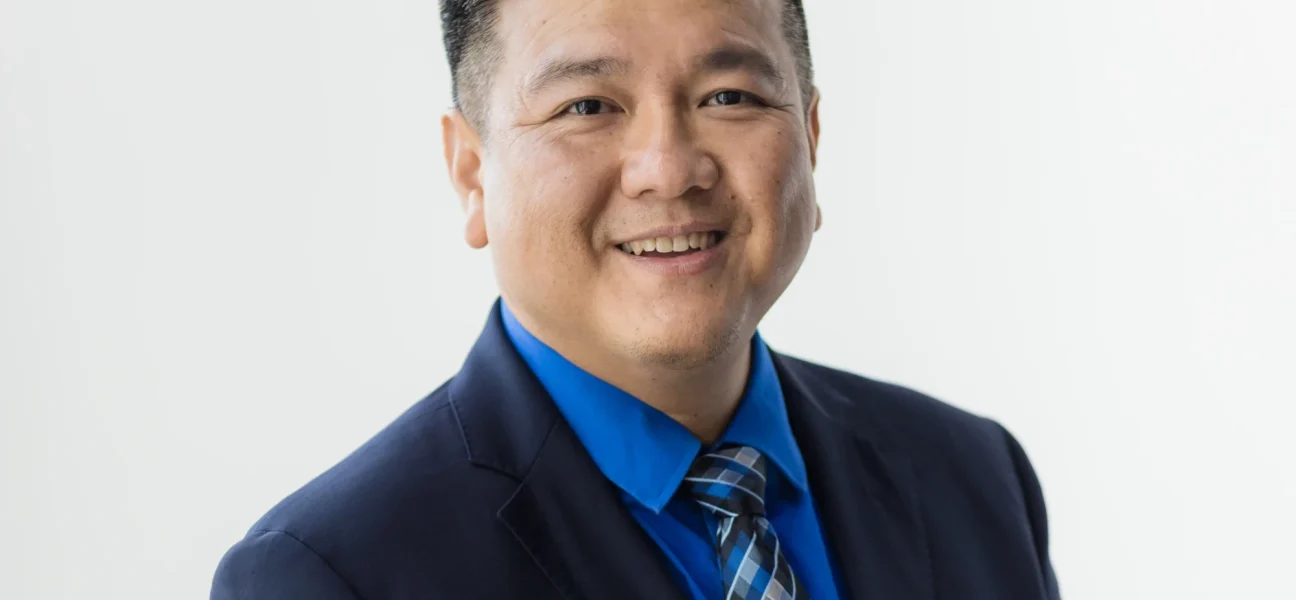 A professional headshot of Naaman Lee, an Asian man with short, styled dark hair, smiling warmly at the camera against a plain white background. He is wearing a dark navy suit jacket, a vibrant blue dress shirt, and a patterned blue and gray tie.