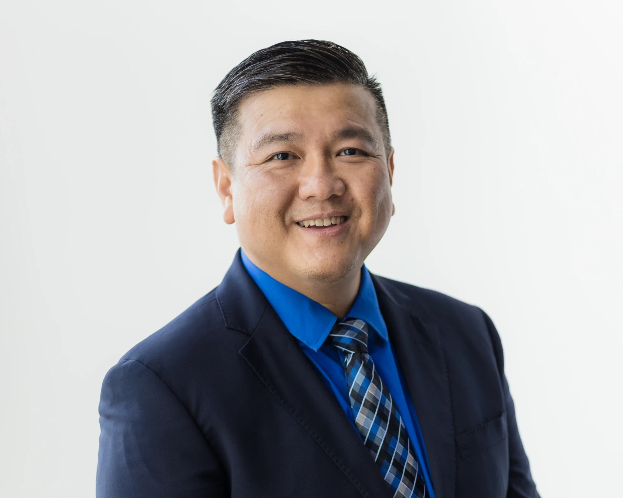 A professional headshot of Naaman Lee, an Asian man with short, styled dark hair, smiling warmly at the camera against a plain white background. He is wearing a dark navy suit jacket, a vibrant blue dress shirt, and a patterned blue and gray tie.