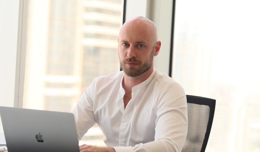 A professional headshot of Peter Ivantsova bald man with a beard wearing a white band-collar shirt, sitting at a desk with a laptop and looking directly at the camera with blurred city high-rises in the background.