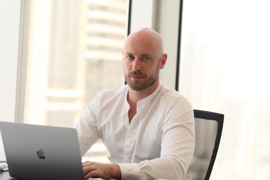 A professional headshot of Peter Ivantsova bald man with a beard wearing a white band-collar shirt, sitting at a desk with a laptop and looking directly at the camera with blurred city high-rises in the background.