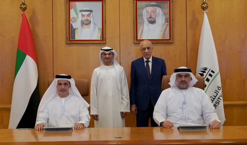 A formal group photo commemorating a meeting between Sharjah Islamic Bank and the University of Sharjah. Two representatives in traditional Emirati attire are seated at a wooden conference table, while two officials stand behind them. The backdrop features a wood-paneled wall with portraits of UAE leadership, flanked by the UAE flag on the left and the University of Sharjah flag on the right.