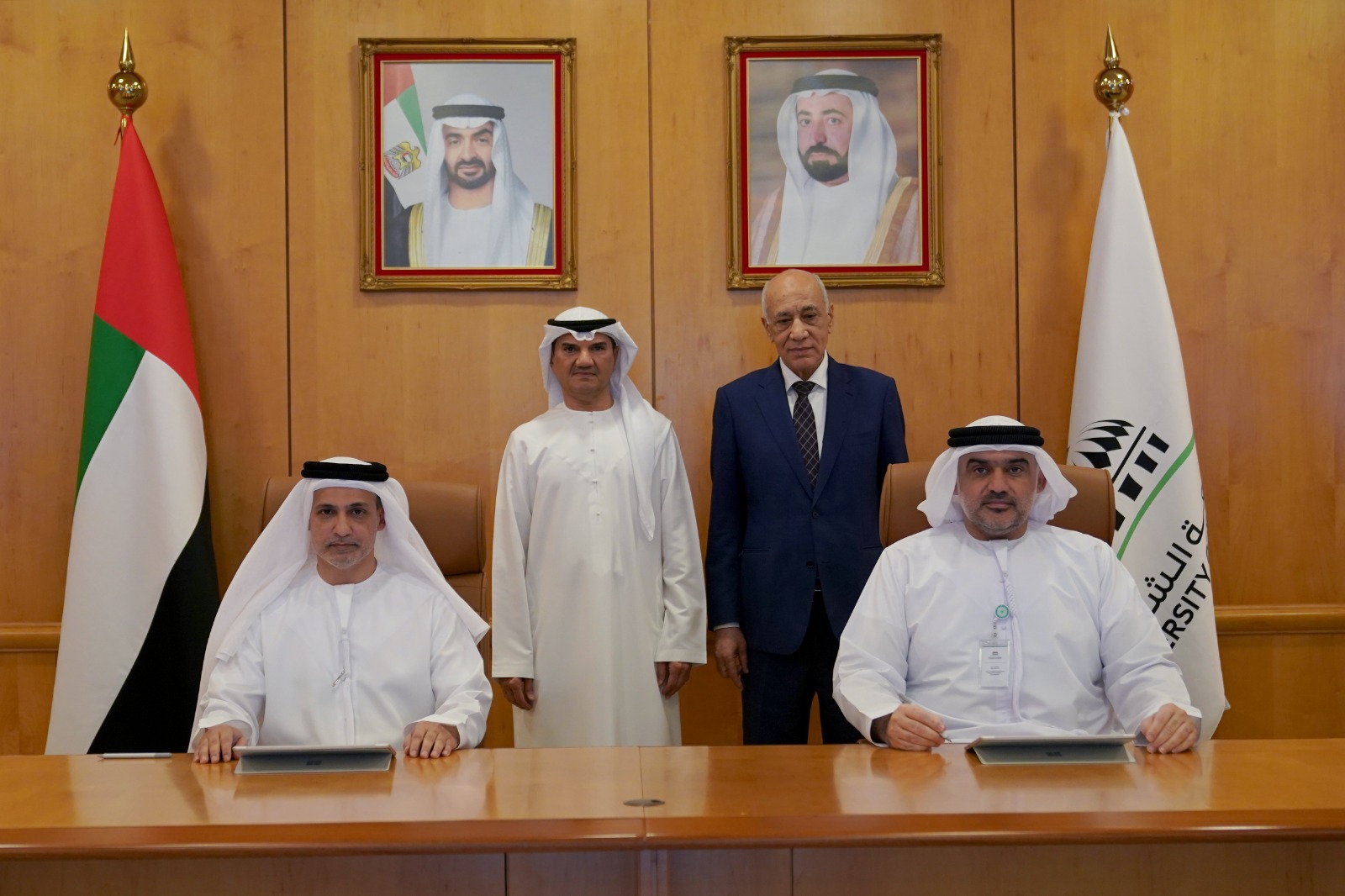 A formal group photo commemorating a meeting between Sharjah Islamic Bank and the University of Sharjah. Two representatives in traditional Emirati attire are seated at a wooden conference table, while two officials stand behind them. The backdrop features a wood-paneled wall with portraits of UAE leadership, flanked by the UAE flag on the left and the University of Sharjah flag on the right.