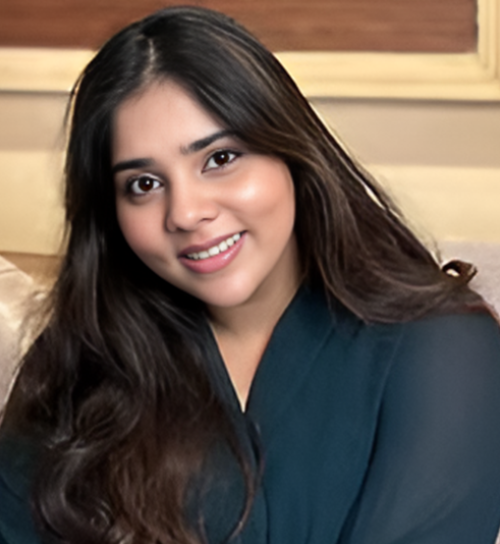 A professional headshot of a woman with long, dark wavy hair smiling warmly at the camera. She is wearing a dark, elegant top and posing against a soft, neutral-toned indoor background.