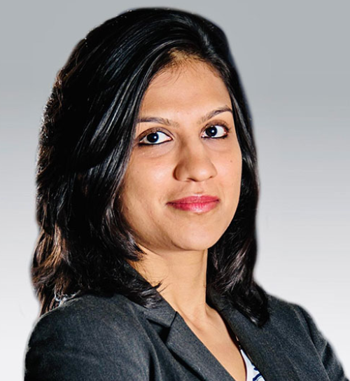 A professional headshot of a woman with shoulder-length layered dark hair looking toward the camera with a subtle smile. She is wearing a dark gray business suit jacket against a neutral gradient background.